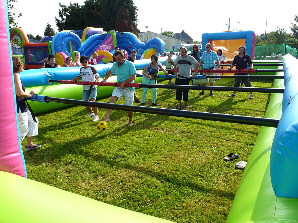 Des participants jouent au babyfoot humain sur un terrain en herbe, utilisant une grande structure gonflable comme terrain, avec un public en arrière-plan.